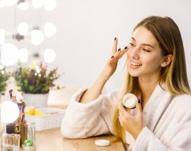 Young beautiful blonde girl cares for face skin with moisturizer in front of mirror.