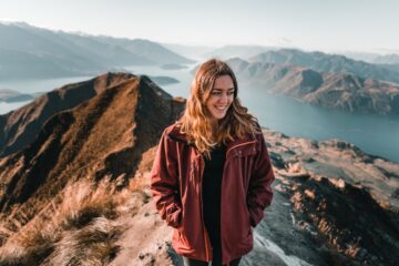blonde brown eyed caucasian young woman on top of a mountain contemplating admiring stunning nature scenery by the big blue lake and rocky mountains on a sunny day, roys peak, new zealand - Travel concept