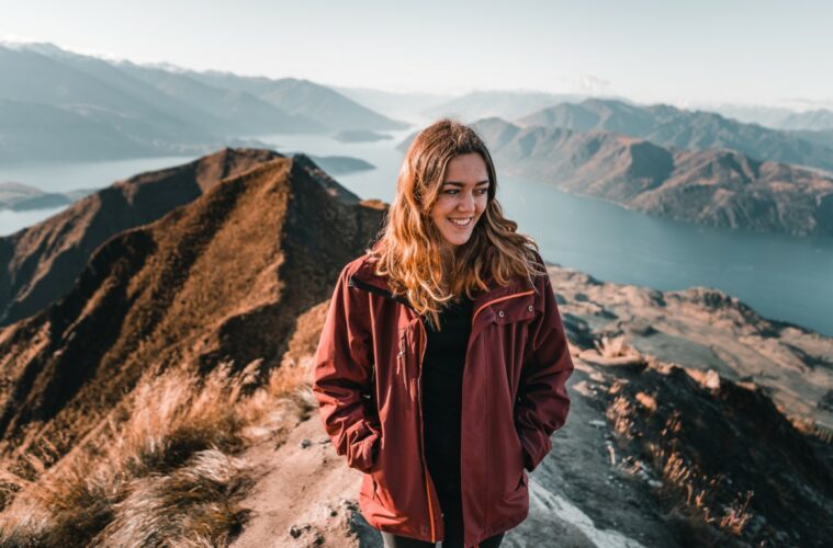 blonde brown eyed caucasian young woman on top of a mountain contemplating admiring stunning nature scenery by the big blue lake and rocky mountains on a sunny day, roys peak, new zealand - Travel concept