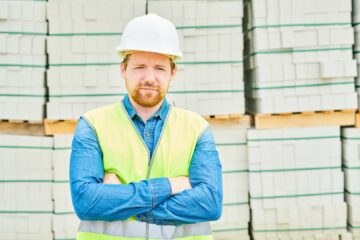 Handsome man in helmet and waistcoat keeping arms crossed and looking at camera while standing on background of stacks of concrete bricks